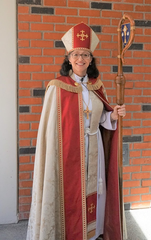 Bishop Megan, wearing a red and cream colored cope and mitre, carrying a wooden crozier.