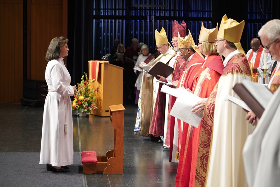 Bishop Megan faces a line of bishops at her consecration. She is wearing a white robe. The bishops are wearing red and white copes and mostly gold mitres. All but one of the other bishops are men.