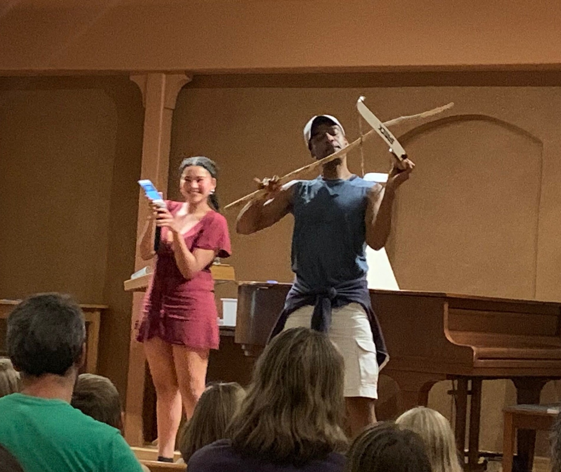 A young woman sings holds up her phone playing music, while her father pretends to play a violin, but its actually a strung bow and walking stick.