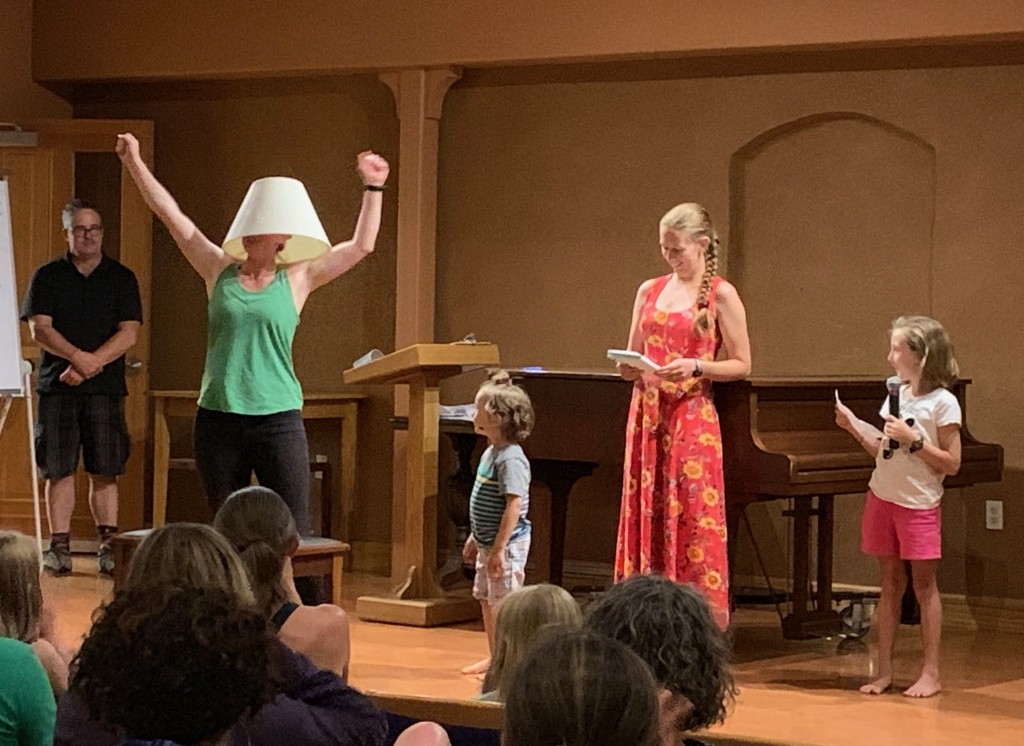 A woman stands on stage with her fists in the air and a lampshade on her head, while two small children and two other adults look on.