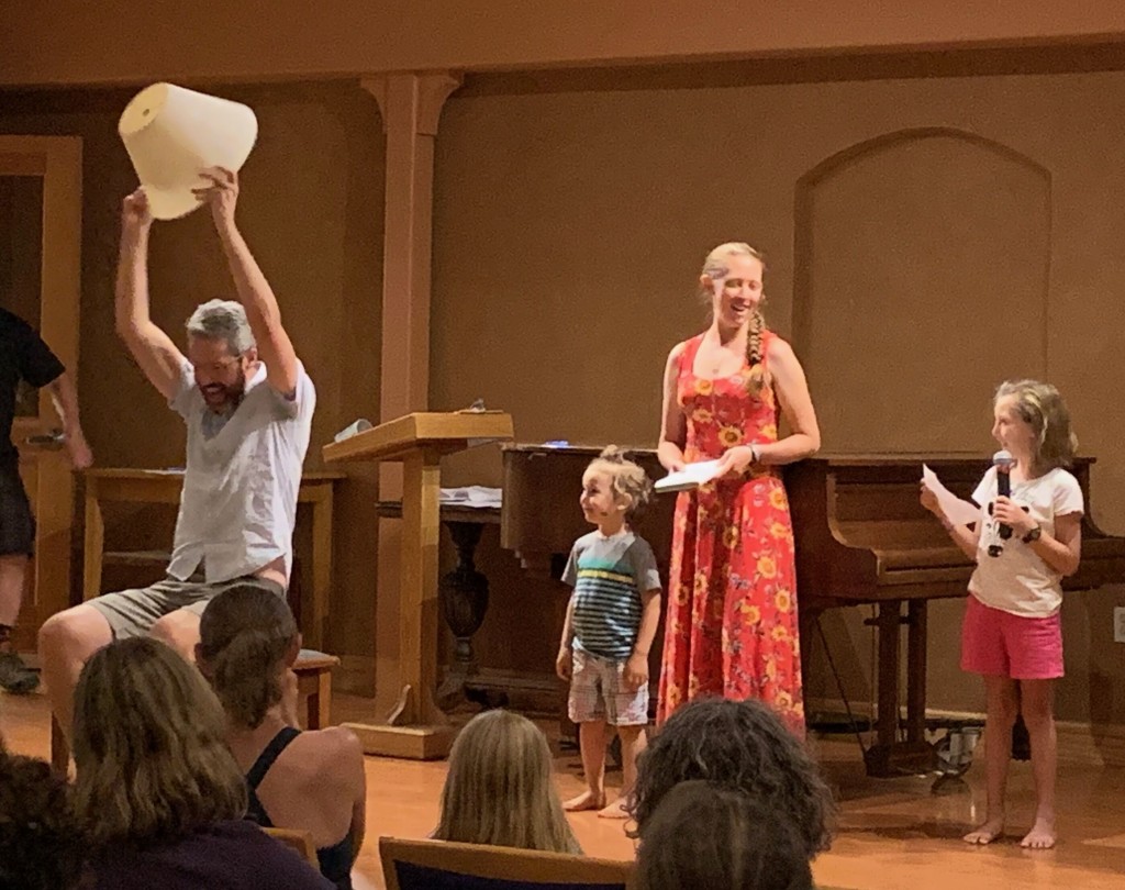 Phil sits on stage holding a lampshade over his head, while two small children and two other adults look on.