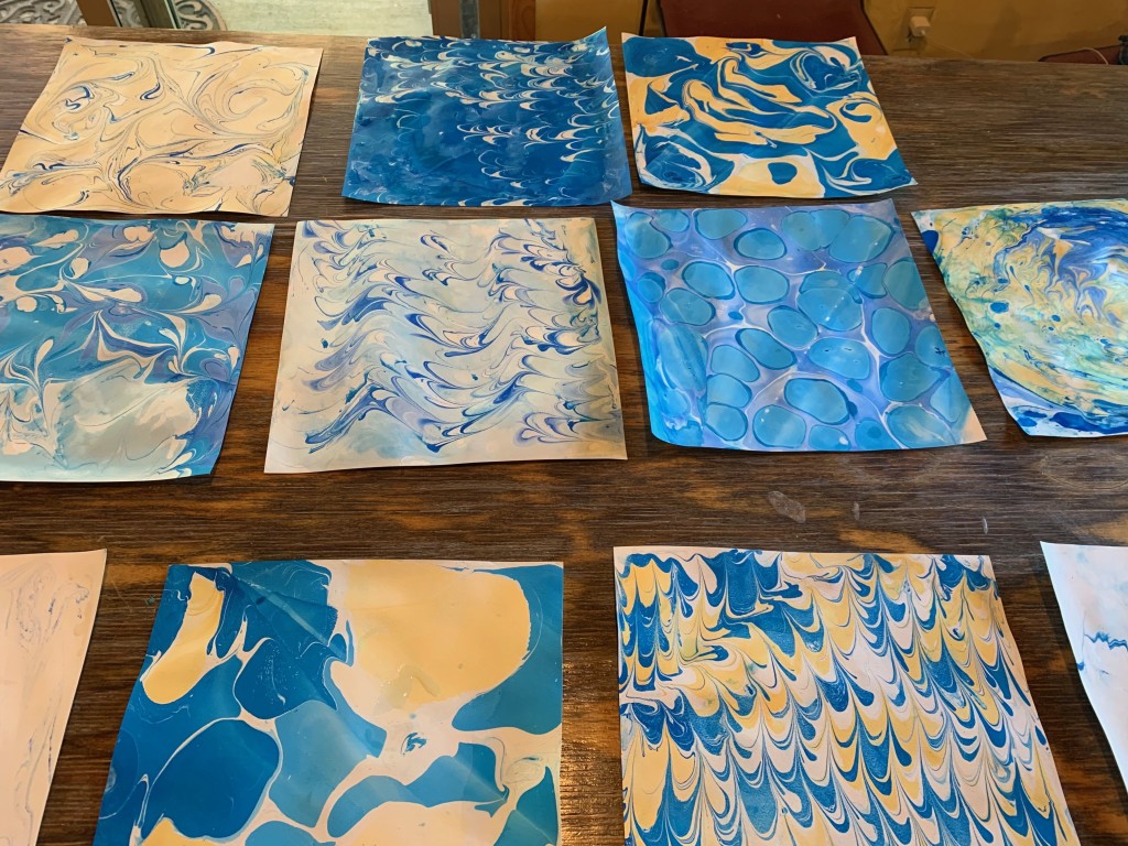Many sheets of marbled paper, in various hues of blue and white, cover a wooden table.